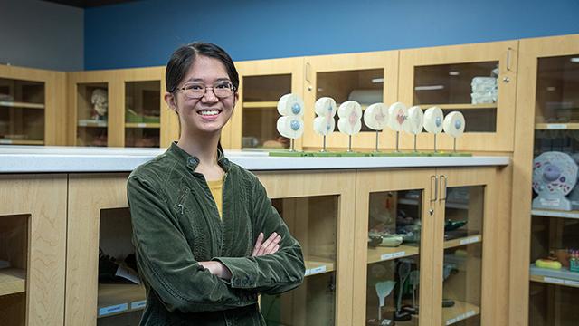 生物学导师凯特琳·赛站在一间科学实验室的房间里，周围的橱柜里放着科学教学工具. To her right is a display showing how a cell divides.  